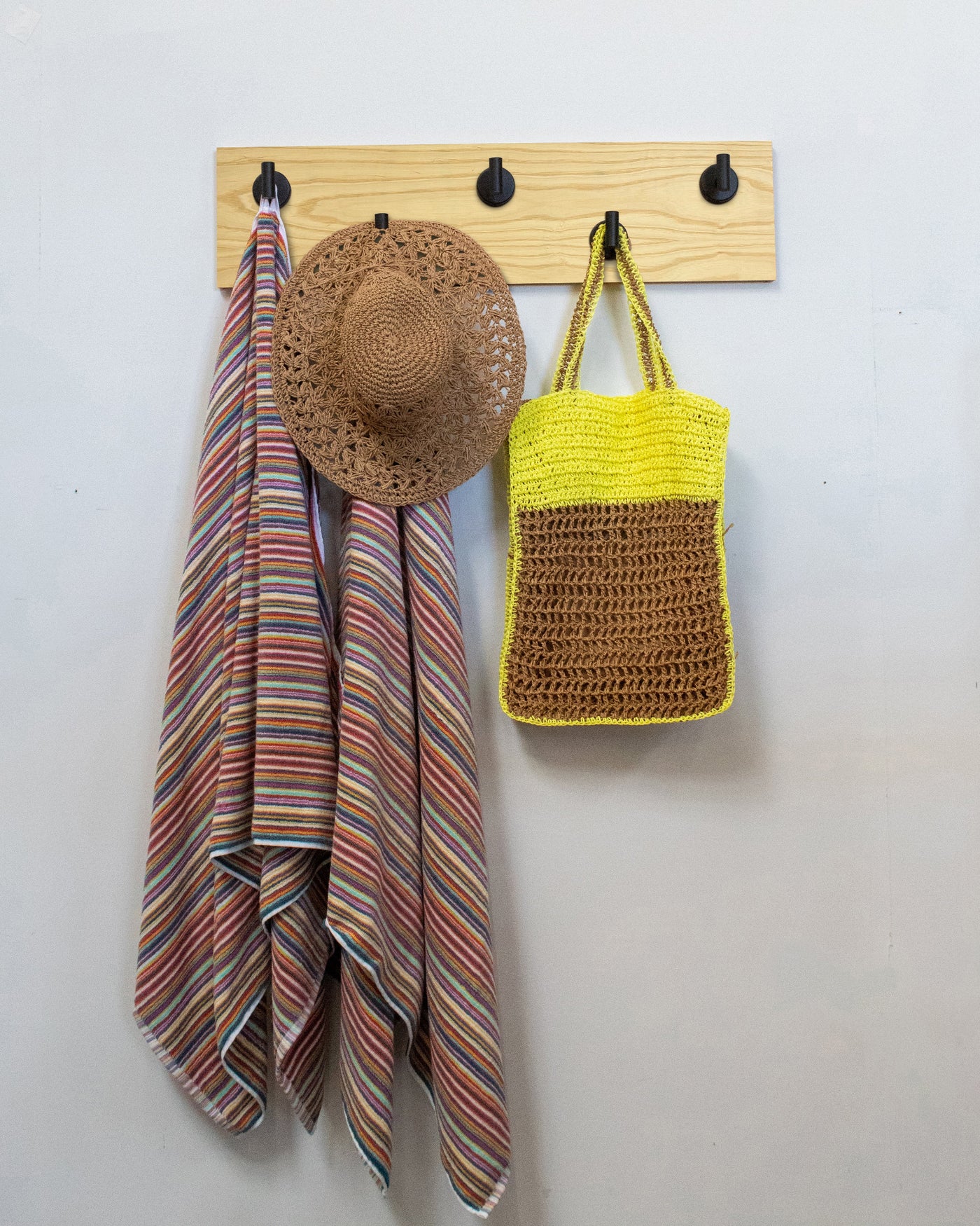 Ash wood wall hook rack with matte black hooks, striped towel, straw hat, and yellow bag on a white wall.