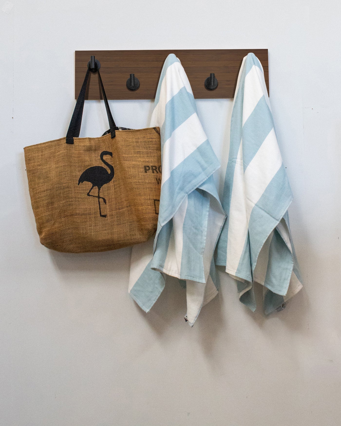 Brown tote bag with flamingo design and two striped towels hanging on a matte black Chocolate Bamboo wood wall hook rack against a white wall.