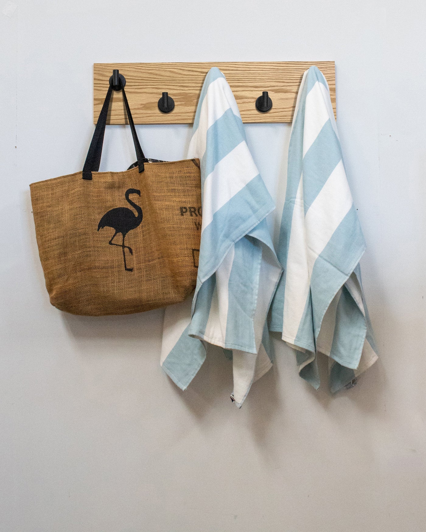 Brown tote bag with flamingo design and two striped towels hanging on a matte black Red Oak wood wall hook rack