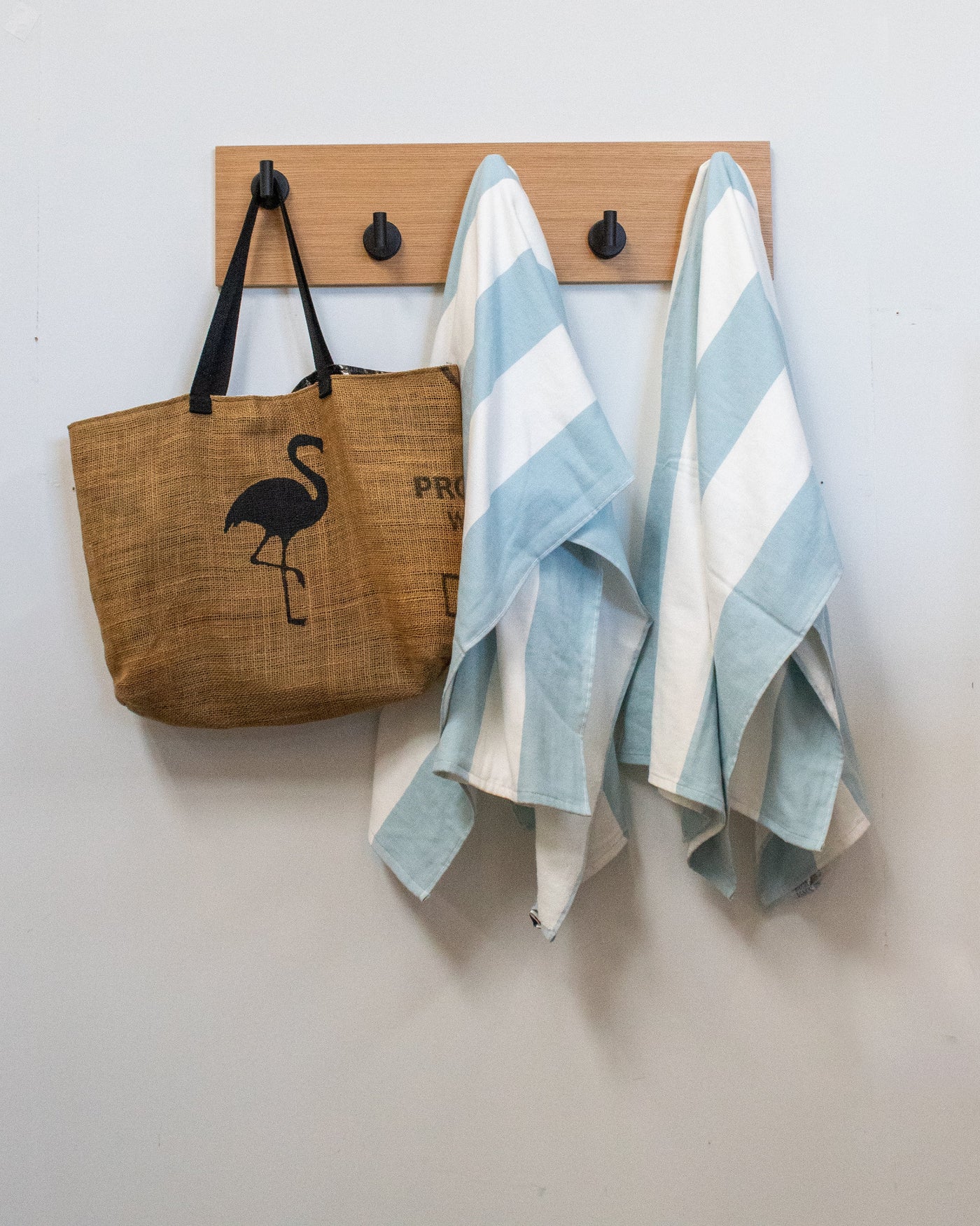 Brown tote bag with flamingo design and two striped towels hanging on a matte black rift white oak wood wall hook rack