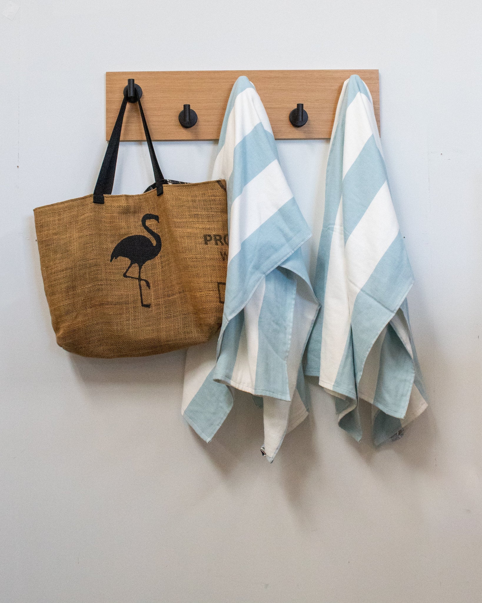 Brown tote bag with flamingo design and two striped towels hanging on a matte black rift white oak wood wall hook rack