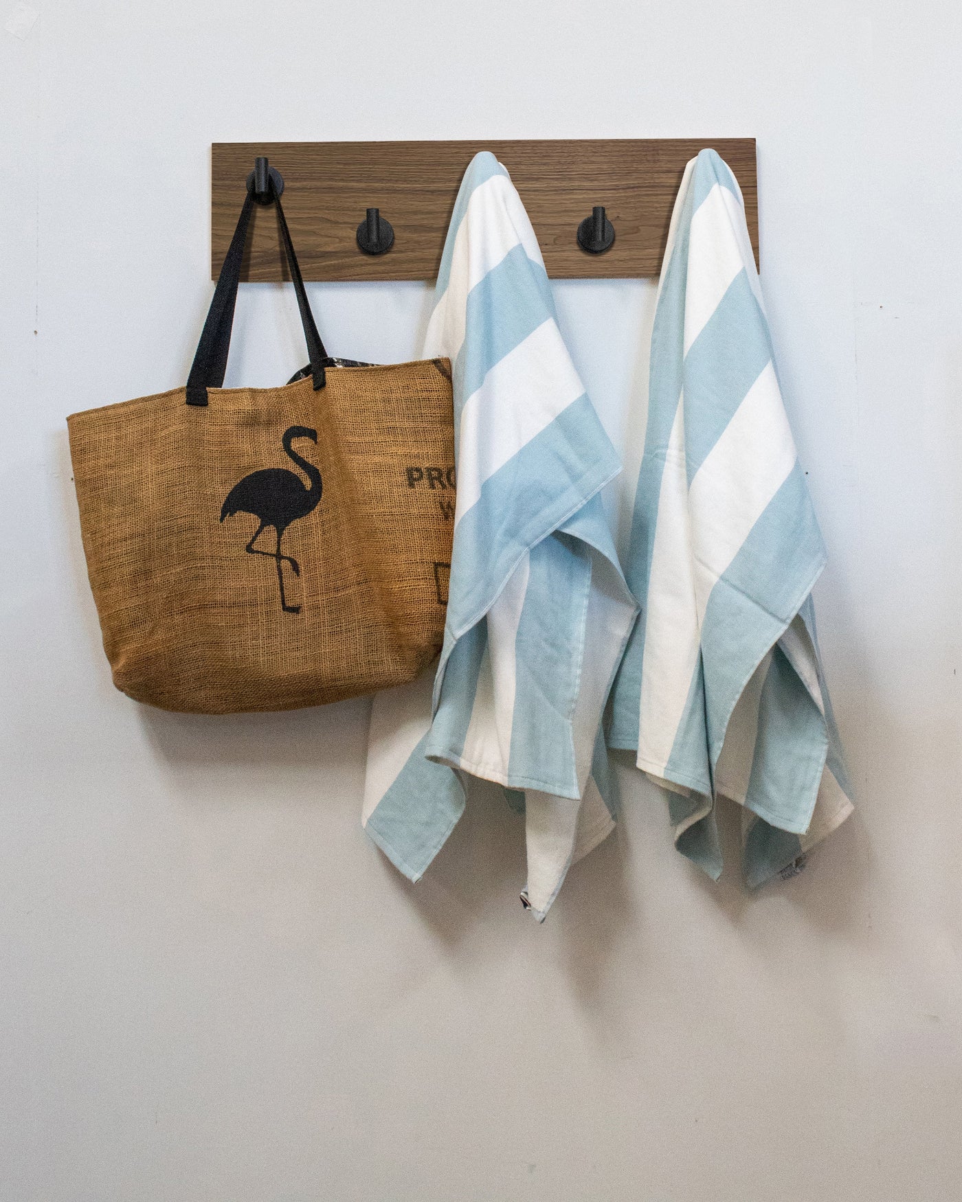 Brown tote bag with flamingo design and two striped towels hanging on a matte black walnut wall hook rack