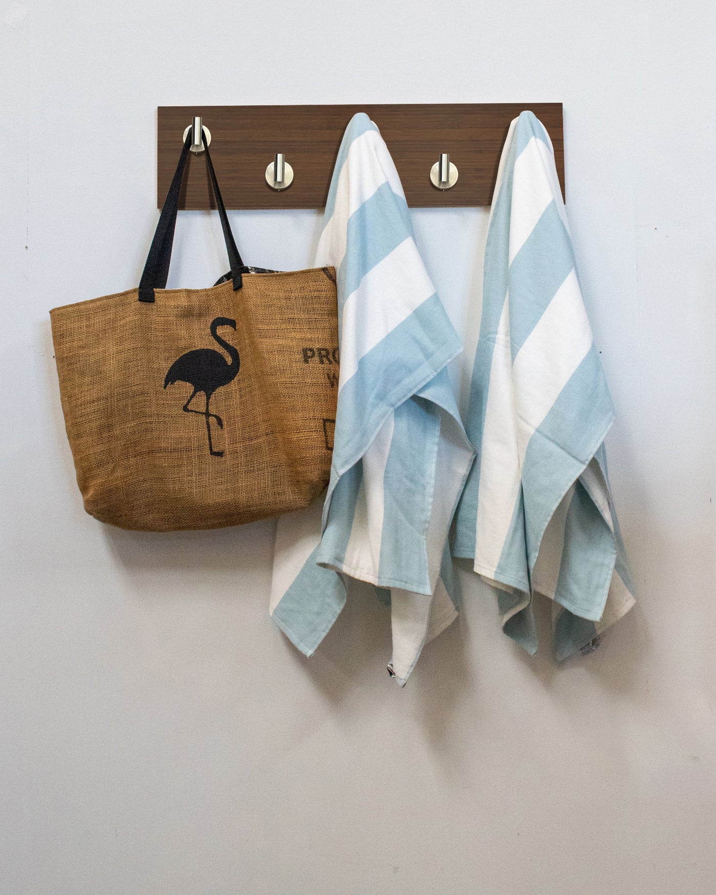 Brown tote bag with flamingo design and two striped towels hanging on a satin nickel Chocolate Bamboo wood wall hook rack against a white wall.