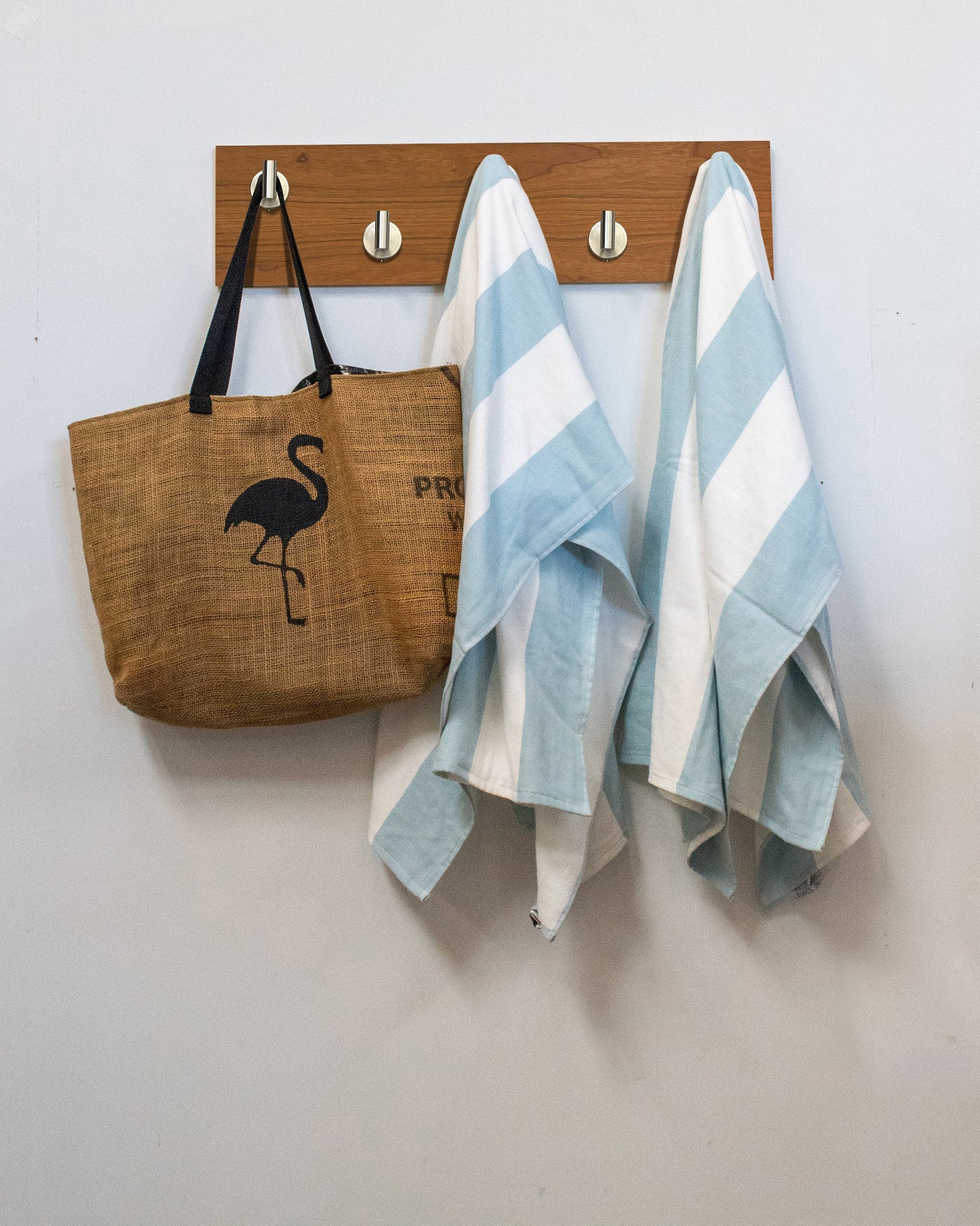 Brown tote bag with flamingo design and two striped towels hanging on a satin nickel cherry wood wall hook rack against a white wall.