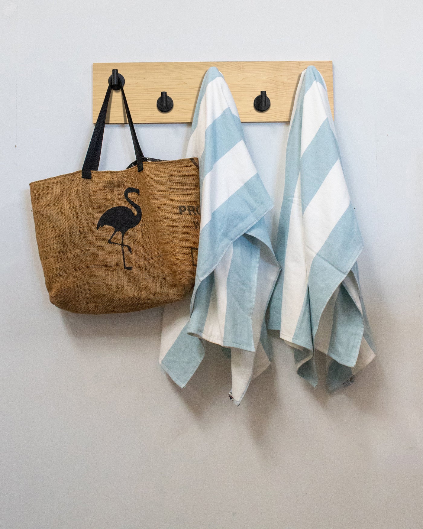 Maple wood wall hook rack with brown tote bag with flamingo design and two striped towels hanging on matte black hooks.