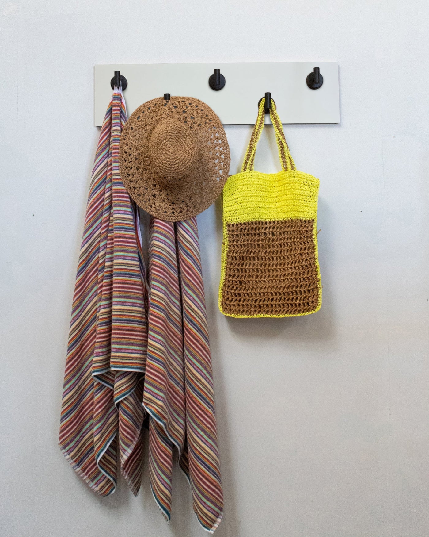 White wood wall hook rack with matte blackl hooks, striped towel, straw hat, and yellow bag on a white wall.